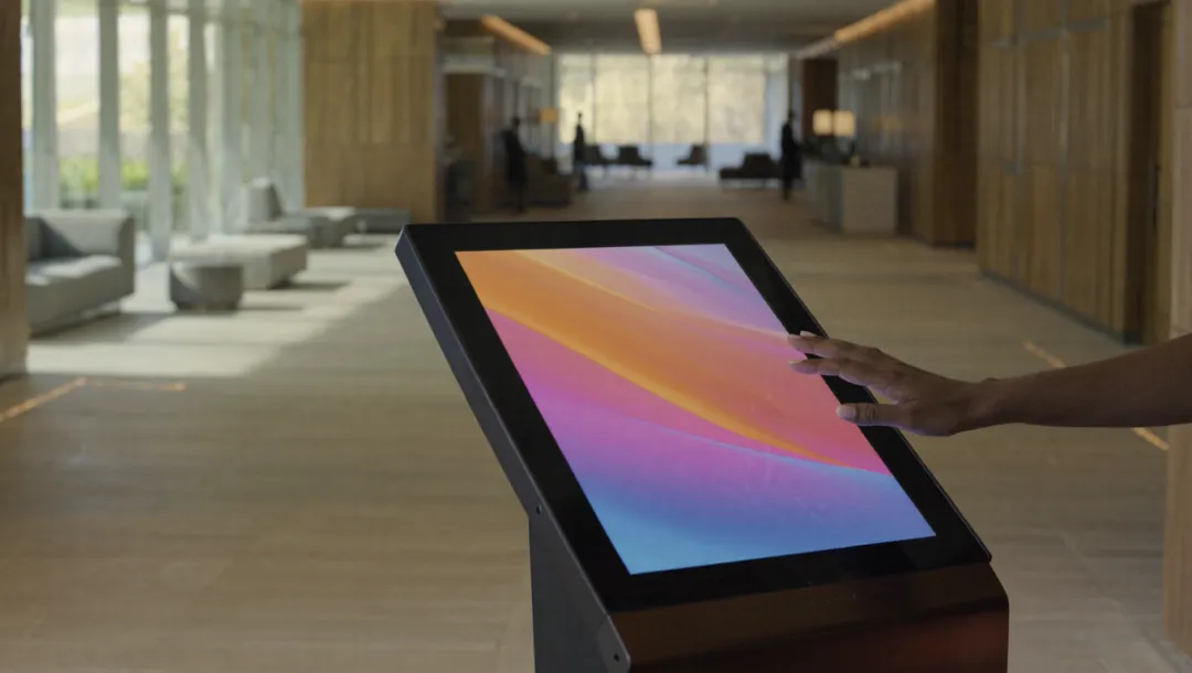 Illustration: a hand touching a tilted touch screen kiosk in a bright lobby